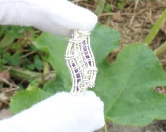 May include: A handcrafted silver pendant with a wire-wrapped design. The pendant features a central row of small, purple gemstones and a clear, central stone. The pendant is held by a gloved hand against a backdrop of green foliage.