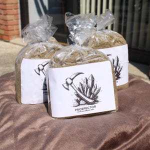 May include: Three clear plastic bags filled with a granular substance, each with a white label featuring a pickaxe and crystals illustration. The text "PROSPECTOR CRYSTAL MINING BAG" is printed on the label. The bags are sitting on a brown surface.