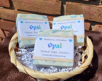 May include: Three rectangular bars of light blue and white swirled soap in a woven basket. Each bar has a white paper label reading Birthstone of October, Opal, Mystical Hope Emotion, Handcrafted Artisan Soap with two small blue heart icons.