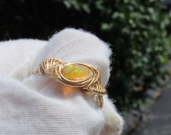 May include: A wire-wrapped ring featuring a central oval-shaped opal gemstone with iridescent green and orange flashes. The band and setting are constructed from thin, coiled gold-toned metal wire that wraps around the stone and forms the ring band.