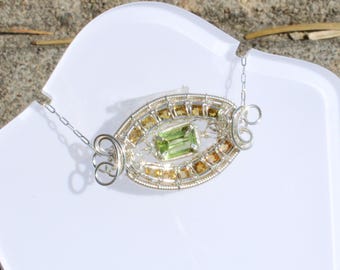 May include: A silver necklace with an oval pendant. The pendant features a central rectangular green gemstone, surrounded by smaller yellow and gold-colored stones. The necklace is displayed on a white surface.
