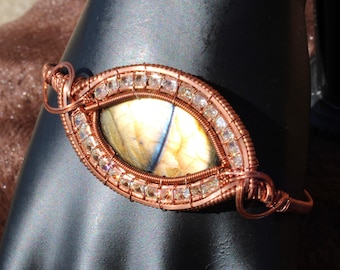 May include: Copper wire-wrapped bracelet featuring a central oval-shaped stone with yellow and blue hues. The stone is surrounded by small, clear crystals and intricate copper wire detailing. The bracelet is displayed on a black surface.