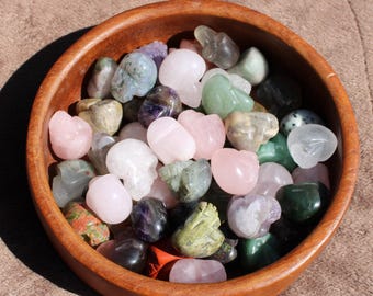 May include: A wooden bowl filled with a variety of polished gemstone skulls in shades of pink, green, purple, and clear. The bowl sits on a brown surface, with the gemstones reflecting light.
