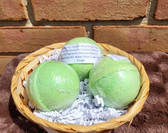 May include: Three spherical light green bath bombs are nestled in a woven wicker basket filled with white paper shred. Each bath bomb features a white adhesive label printed with ingredients including Sodium Bicarbonate, Citric Acid, and Blue 1 Lake.