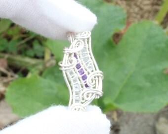 May include: A handcrafted silver wire pendant with a central design of clear and purple stones. The pendant is held by a gloved hand against a backdrop of green foliage. The wirework forms an intricate, open pattern.