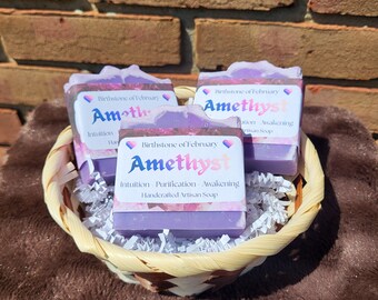 May include: Three rectangular bars of purple soap are nestled in a woven basket with white paper shred filler. Each bar features a label that reads Birthstone of February Amethyst Intuition Purification Awakening Handcrafted Artisan Soap.