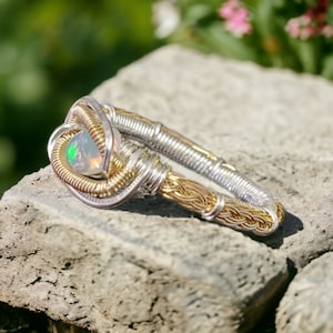 May include: A silver and gold wire wrapped ring with an iridescent opal gemstone. The ring is sitting on a rough, gray rock.