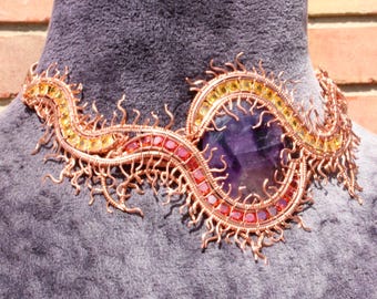 May include: A handcrafted copper wire necklace with a large, oval, purple gemstone at the center. The necklace features a wave-like design with embedded red and yellow gemstones. The intricate wirework has a textured, spiky appearance.
