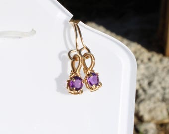 May include: A pair of gold-colored wire-wrapped earrings. Each earring features a round, vibrant purple gemstone. The earrings are suspended from a simple hook design, showcasing the gemstones' color and intricate wirework.