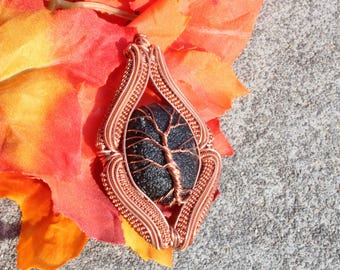May include: A copper wire-wrapped pendant featuring a dark, textured stone with a wire tree design. The pendant is set against a backdrop of orange and red autumn leaves, creating a natural, earthy aesthetic.