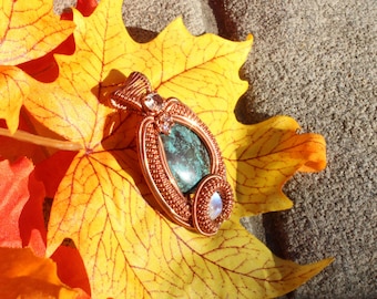 May include: A handcrafted copper wire pendant with a teal stone center and a smaller moonstone accent. The pendant is displayed on a vibrant yellow leaf, with other leaves in the background. The pendant is an example of artisan jewelry.