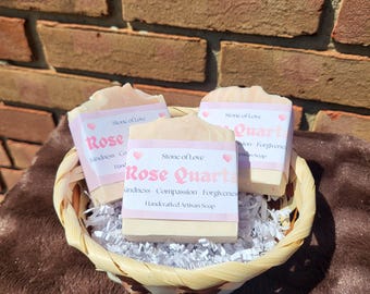 May include: Three rectangular bars of pale pink handcrafted artisan soap nestled in a woven basket with white paper shred. Each soap features a white label with pink text reading Stone of Love, Rose Quartz, Kindness, Compassion, Forgiveness.