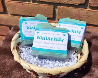 May include: Three rectangular bars of teal and green marbled soap are nestled in a woven basket with white paper shred. Each bar features a white label with the text Stone of Harmony, Malachite, Strength, Abundance, Cleansing, Handcrafted Artisan Soap.