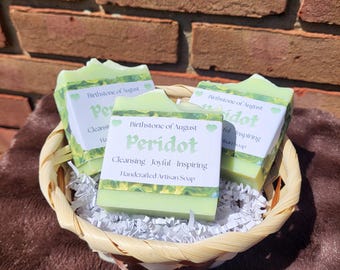 May include: Three rectangular bars of light green soap arranged in a woven basket filled with white paper shreds. Each soap features a white label with the text Birthstone of August, Peridot, Cleansing, Joyful, Inspiring, Handcrafted Artisan Soap.