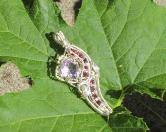 May include: A handcrafted silver pendant with a central light purple gemstone and smaller red gemstones. The pendant has a detailed wire-wrapped design and is displayed on a large green leaf, showcasing its intricate craftsmanship and natural setting.