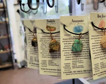 May include: Three necklaces with different colored stones and metal wire wrapped around them. The stones are green, brown, and pink. The necklaces are labeled with the names of the stones: Unakite, Amazonite, and Rose Quartz. The labels also include information about the stones' properties and uses.