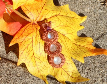 May include: A copper wire-wrapped pendant with three moonstone cabochons and a small purple gemstone. The pendant is displayed on a large yellow leaf with orange and red accents, set against a gray stone surface.