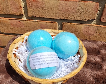 May include: Three spherical turquoise bath bombs nestled in a woven wicker basket filled with white paper shred. One bath bomb features a white label with the text: Turquoise - Sodium Bicarbonate, Citric Acid, Magnesium Sulfate, Essential Oil/Fragrance, Pomace Olive Oil, Polysorbate 80, Water, Blue 1.