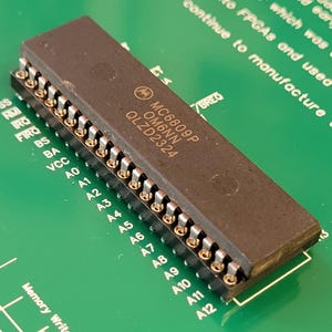 May include: A close-up of a rectangular, dark brown integrated circuit chip with gold-coloured pins. The chip is labelled "MC6809P OM6NN QLZD2324" and is set against a green circuit board with text.