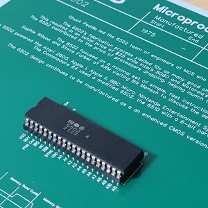 May include: A black MOS 6502 microprocessor chip on a green background with white text. The chip has multiple pins and the text "6502 2122 A". The background text describes the chip's history and use in early computers.