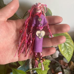 May include: A purple fabric doll with pink and white yarn hair, a heart charm with a key, and a heart charm with an angel. The doll has gold bells on its feet.