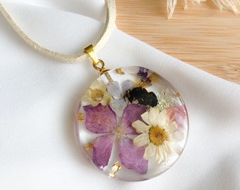 May include: A round, clear resin pendant necklace with a white suede cord. The pendant features pressed purple and white flowers, gold leaf, and a small black bee.