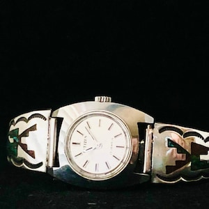 Könnte beinhalten: Eine silberne Uhr mit weißem Zifferblatt und schwarzen Zeigern. Das Armband der Uhr ist mit Türkis und Silber-Intarsien in einem südwestlichen Design verziert.