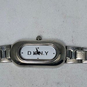 Puede incluir: Un reloj plateado con una esfera blanca y la inscripci&oacute;n DKNY en negro. El reloj tiene una pulsera de metal plateado.