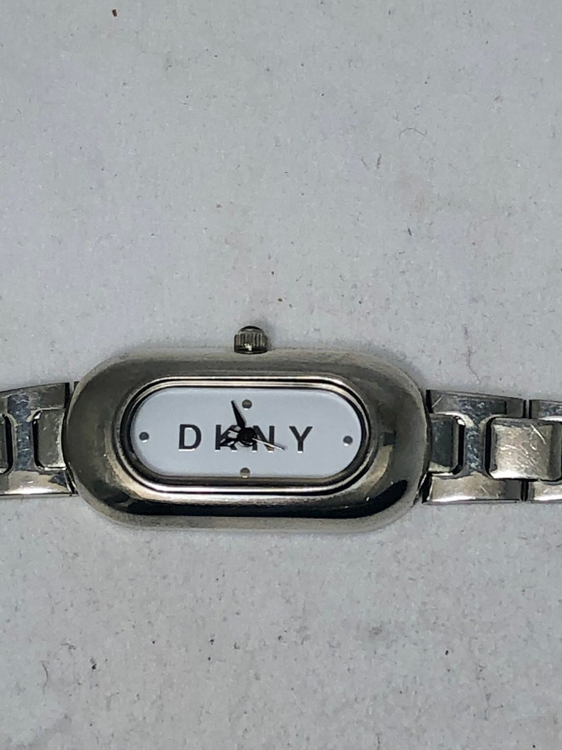Puede incluir: Un reloj plateado con una esfera blanca y la inscripci&oacute;n DKNY en negro. El reloj tiene una pulsera de metal plateado.