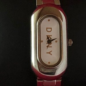 Puede incluir: Un reloj de pulsera plateado y rojo con una esfera blanca y la palabra "DKNY" impresa en letras doradas.