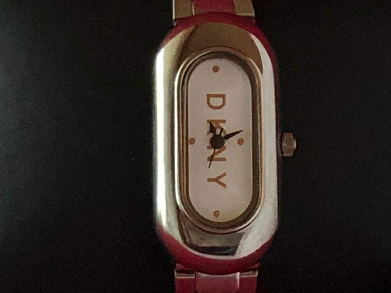 Puede incluir: Un reloj de pulsera plateado y rojo con una esfera blanca y la palabra "DKNY" impresa en letras doradas.