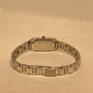 Puede incluir: Un reloj plateado con una esfera rectangular y una correa de metal plateado.