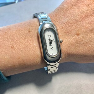 Puede incluir: Un reloj plateado con una esfera blanca y una correa plateada. La esfera del reloj tiene la palabra "DKNY" impresa.