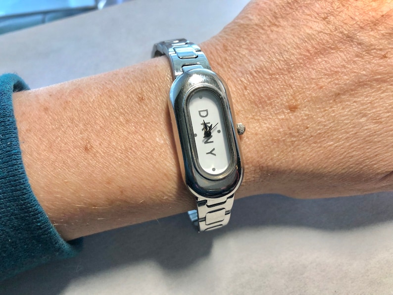 Puede incluir: Un reloj plateado con una esfera blanca y una correa plateada. La esfera del reloj tiene la palabra "DKNY" impresa.