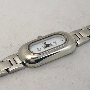 Puede incluir: Un reloj plateado con una esfera blanca y una correa plateada. La esfera del reloj tiene la palabra "DKNY" impresa.