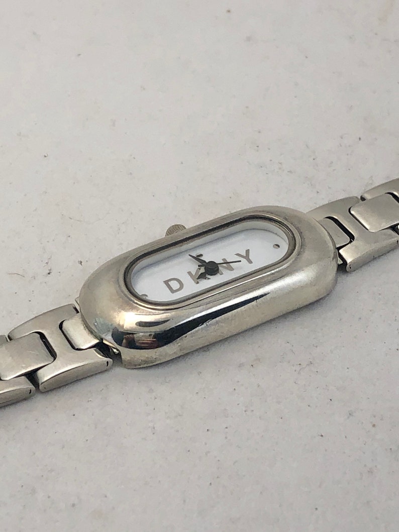 Puede incluir: Un reloj plateado con una esfera blanca y una correa plateada. La esfera del reloj tiene la palabra "DKNY" impresa.