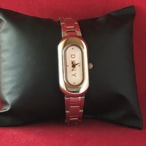 Puede incluir: Un reloj plateado con una esfera rectangular y una esfera blanca. El reloj tiene una correa plateada y la palabra "DKNY" est&aacute; impresa en la esfera.