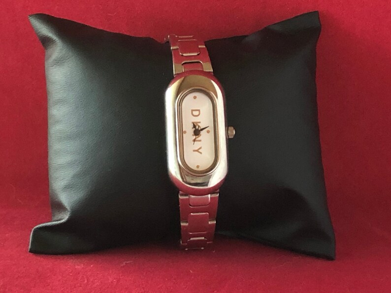 Puede incluir: Un reloj plateado con una esfera rectangular y una esfera blanca. El reloj tiene una correa plateada y la palabra "DKNY" est&aacute; impresa en la esfera.