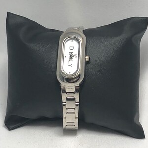 Puede incluir: Un reloj plateado con una esfera blanca y la palabra "DKNY" impresa en &eacute;l. El reloj tiene una correa plateada y una esfera rectangular.