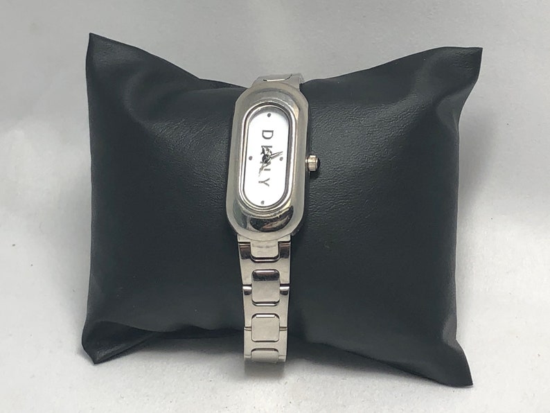 Puede incluir: Un reloj plateado con una esfera blanca y la palabra "DKNY" impresa en &eacute;l. El reloj tiene una correa plateada y una esfera rectangular.