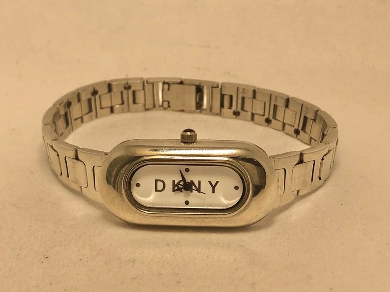 Puede incluir: Un reloj plateado con una esfera blanca y agujas negras. El reloj tiene una esfera rectangular con bordes redondeados y el nombre de la marca "DKNY" impreso en la esfera.