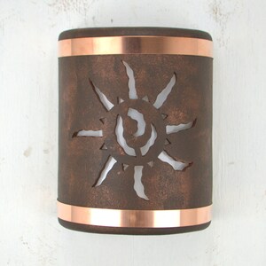 May include: A copper wall sconce with a cutout design of a sun. The sconce has a rustic brown finish and is accented with copper bands.