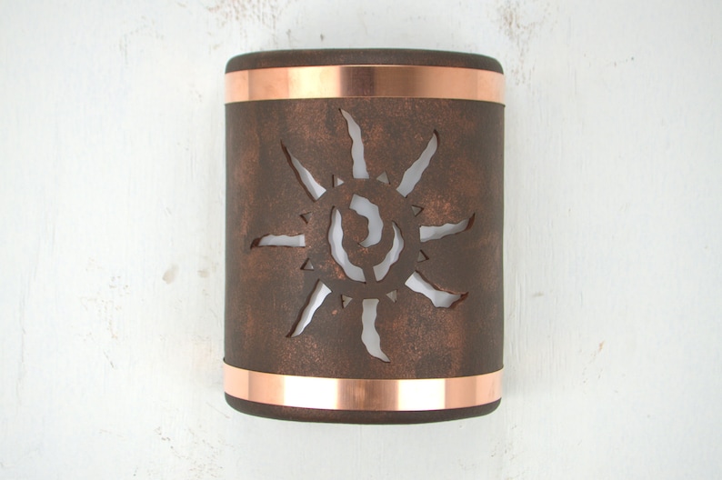 May include: A copper wall sconce with a cutout design of a sun. The sconce has a rustic brown finish and is accented with copper bands.