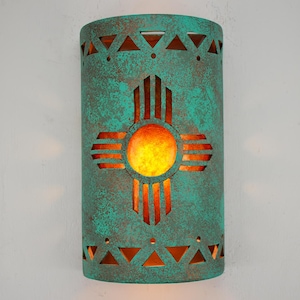 May include: A turquoise green metal wall sconce with a cut-out design of a Zia symbol. The sconce has a warm orange glow emanating from the light source.