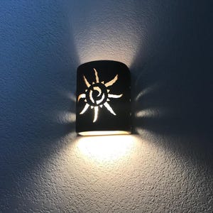 May include: A brown ceramic wall sconce with a cut-out sun design. The sconce is lit and casting a warm glow on the wall.
