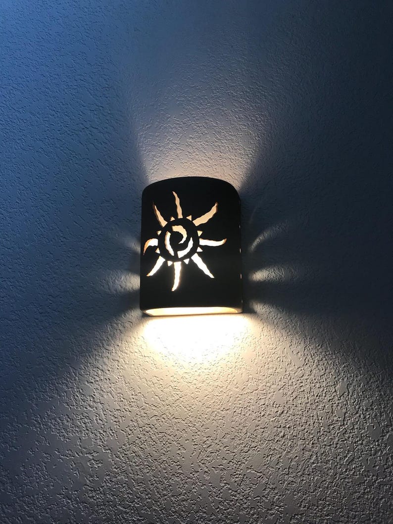 May include: A brown ceramic wall sconce with a cut-out sun design. The sconce is lit and casting a warm glow on the wall.