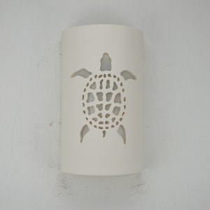 May include: White ceramic wall sconce with a cutout design of a sea turtle. The sconce is a cylinder shape and the turtle is centered on the front.