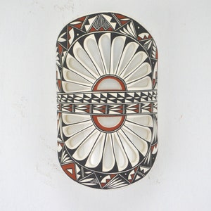 May include: A white ceramic wall hanging with a geometric design in black and red. The design features a central circle with radiating lines and a band of triangles around the center.
