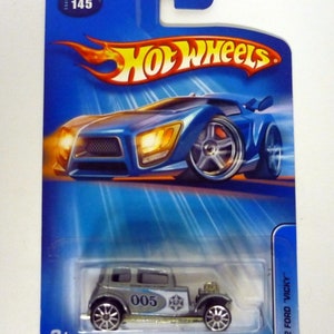 Könnte beinhalten: Eine blaue Hot Wheels-Verpackung für ein Spielzeugauto aus Metallguss mit einem silbernen 1932 Ford Vicky-Spielzeugauto im Inneren. Das Auto trägt die Nummer "005" an der Seite. Die Verpackung zeigt das Hot Wheels-Logo und den Text "1932 Ford Vicky".
