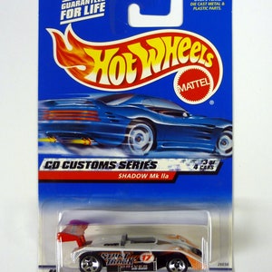 Puede incluir: Un coche de juguete Hot Wheels de metal fundido a presión de color blanco con detalles rojos y negros. El coche está etiquetado como "Shadow Mk lla" y "CD Customs Series". El coche es parte de una serie de 4 coches y es el número 3 de 4. El coche está en una tarjeta azul y blanca con el logotipo de Hot Wheels.